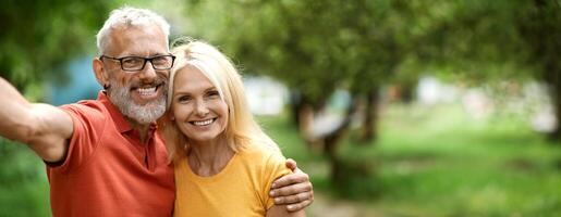 A joyful older couple stands close together, smiling widely while embracing and taking selfie in a vibrant park filled with green trees and soft grass. The sun shines brightly, copy space photo