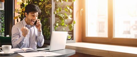 Unbelievable success. Excited millennial businessman in cafe with open laptop, looking at computer screen and talking on phone, empty space photo