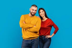 A man and woman standing together, smiling, and posing in front of the camera. They are dressed in casual attire and appear relaxed, blue background photo