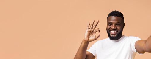 A smiling Black man with a beard and a white t-shirt is making the okay sign with his hand, looking directly at the camera. He is standing against a light brown background, copy space photo