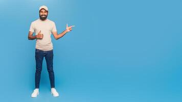 A casually dressed man stands against a vibrant blue backdrop, smiling while giving a thumbs up with one hand and pointing with the other, copy space photo