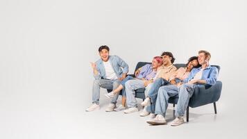 A group of five friends are enjoying each others company, laughing and smiling. The diverse group of young adults are casually dressed in denim and sitting on a couch in a light studio space. photo
