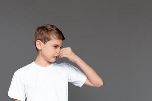 A young boy wearing a white t-shirt is pinching his nose, an expression of disgust on his face as he reacts to an unpleasant smell, isolated on a grey background photo