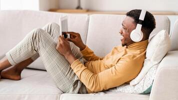 Music App. Cheerful Black Man In Headphones Using Digital Tablet Relaxing On Sofa At Home. Selective Focus photo