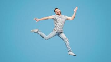 Funny millennial European guy jumping in air and fooling on blue studio background, full length, empty space photo