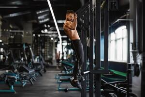 Muscular guy with naked torso pulling up on horizontal bar in gym, empty space photo