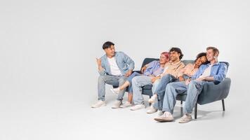 A group of diverse friends enjoy each others company while sitting on a couch. One friend is seated slightly apart from the others, leaning forward with his arms crossed as he speaks, copy space photo