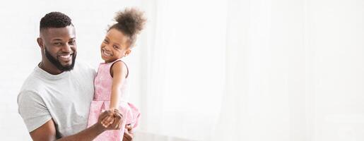A joyful black father holds his smiling daughter in his arms as they dance playfully. Sunlight filters through the bright room, illuminating their happiness and creating a warm atmosphere. photo