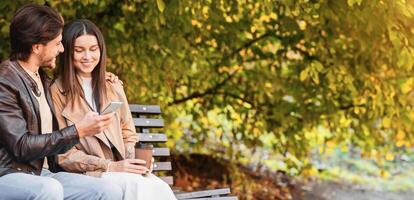 Man and woman sitting on bench and watching photos on mobile phone, spending time together in park, copy space