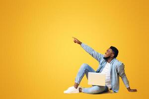 Look there. African american guy sitting on floor with laptop and pointing aside at copy space on yellow background photo