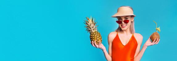 A young woman is wearing an orange swimsuit and a straw hat, and holding a pineapple in one hand and a coconut with a straw in the other. She is smiling and looking directly at the camera. photo