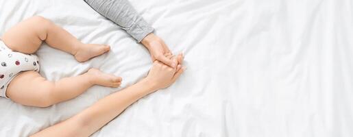 A parent gently holds the tiny hand of their infant while both rest on a soft, light-colored bedspread. The atmosphere is calm and nurturing, reflecting a deep bond between them, cropped photo