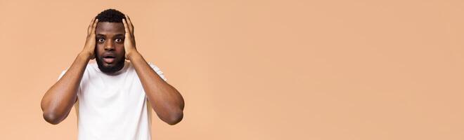 A shocked young Black man is shown in this image. He is wearing a white t-shirt and has his hands on his head as if he is surprised or in disbelief, panorama with copy space photo