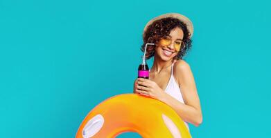 Black woman with curly hair smiles brightly as she poses in front of a blue backdrop. She is holding a soda with a straw and an inflatable ring, suggesting a day of fun and relaxation in the sun. photo