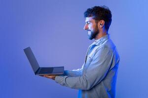 A man with a beard is standing while holding a laptop in his hands. He appears to be looking at the screen intently, possibly working or browsing the internet. photo