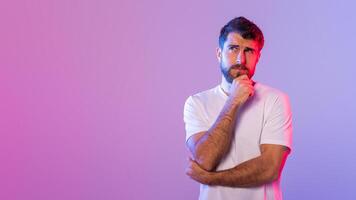 A young man dressed in a white t-shirt stands against a vivid gradient background, appearing thoughtful as he rests a hand on his chin in contemplation, copy space photo