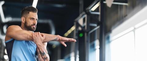 A man performs arm stretches in a modern gym, focusing on flexibility and fitness as sunlight streams through large windows, copy space photo