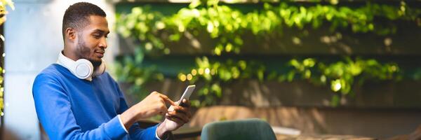 Technology Concept. Side view of young african american student in headphones using smartphone sitting at modern cafe, panorama with copy space photo