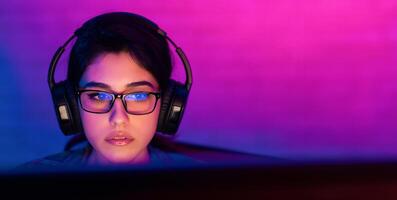 A young woman wearing glasses and headphones is focused on a computer screen. The room is lit with a soft, pink and blue gradient, creating a moody and atmospheric feel, copy space photo