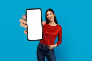 A woman wearing a red shirt is holding up a cell phone with blank screen in her hand, the screen is visible. She appears to be engaging with the device, possibly taking a photo or sending a message.