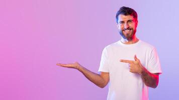 A cheerful man with a beard, wearing a white t-shirt, is pointing to his left hand while standing against a vibrant, colorful gradient background, copy space photo