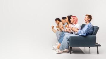 Five diverse young friends are sitting on a couch in a studio setting. They are all looking off to the side, cheering excitedly. They are likely watching a sporting event or a game show, copy space photo