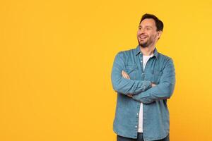 A man with a beard and short brown hair wears a blue denim shirt with a white undershirt. He stands in front of a yellow background, smiling, with his arms crossed. He is looking at copy space photo