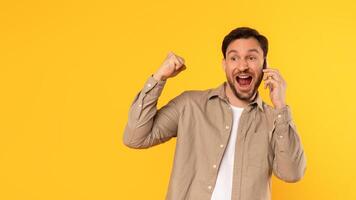 A man with a beard is talking on his phone with an excited expression. He is standing in front of a yellow background and has raised his right arm in the air, copy space photo
