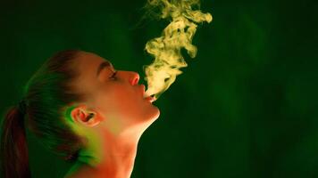 A woman is standing in front of a plain green background, smoking a cigarette. She is blowing smoke into the air, copy space photo
