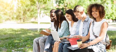 Group of students studying together outdoors, preparing for lectures and classes while sitting on bench in campus, black girl smiling at camera photo
