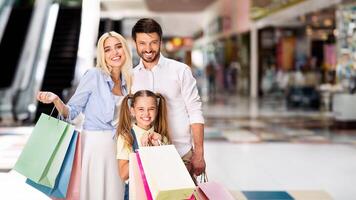A young family, a mother, father, and daughter, are standing in a large mall. They are all smiling happily and holding shopping bags, copy space photo