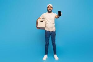 Indian delivery man is standing and smiling while holding a cardboard box in one hand and displaying a smartphone in the other photo