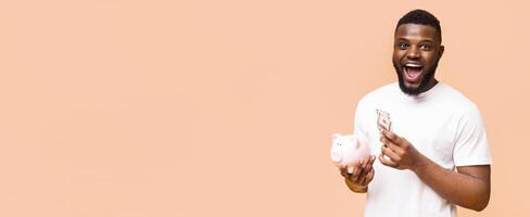 Black man in a white t-shirt is holding cash and a piggy bank in front of a peach-colored background. He is looking at the camera and has a surprised expression on his face, copy space photo