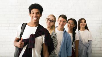 Five cheerful teenagers, three girls and two boys, are seen lined up against a white brick wall background. The group appears to be friends photo