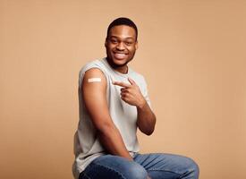 Vaccination Against Covid-19. Joyful Vaccinated African Guy Pointing Finger At Adhesive Plaster Bandage On His Arm After Coronavirus Antiviral Injection Sitting On Beige Background. I've Got Vaccine photo