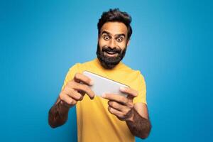A joyful Indian man wearing a yellow shirt is enthusiastically playing a mobile game. He is set against a vibrant blue background, fully immersed in the game. photo
