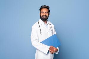 A cheerful Indian doctor is seen holding a blue folder while standing against a plain blue background. He is wearing a white lab coat and a stethoscope around his neck photo