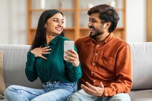 Indian man and woman are seated on a couch, both engrossed in a cell phone they are holding. They appear focused on the screen, perhaps watching a or browsing social media together. photo