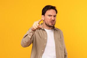A man is shown in a studio, wearing a light brown button-down shirt over a white t-shirt, looking directly at the camera with a furious expression and his index finger pointed out photo