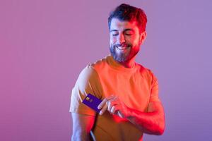 A man is standing holding a credit card in his right hand. He appears happy and relaxed, studio background with neon light photo