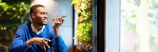 Voice Search. Happy black man using voice assistant on mobile phone or calling on smartphone at cafe, copy space, web-banner photo