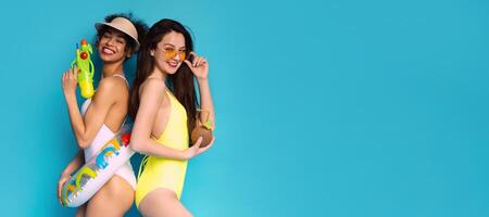 Two young women are posing in their swimsuits in front of a blue background. They both have smiling expressions, copy space photo