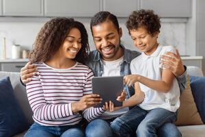 African American man, woman, and child are seated on a couch, engrossed in what appears to be a tablet device. They are focused on the screen, with expressions of concentration and curiosity. photo