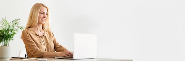 Blonde millennial business lady sitting at workplace, working on laptop at cozy office, chatting with clients, sending emails, panorama with copy space photo