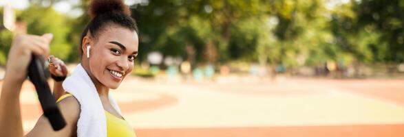 Portrait of smiling African American woman doing workout training using equipment for body stretching exercise outside. Attractive lady enjoying outdoor activity, panorama with copy space photo