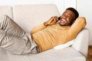 Phone Call. Happy Afro Man Talking On Cellphone Lying On Couch At Home. photo