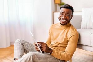 Positive Afro Man Using Tablet Computer Smiling To Camera Sitting On Floor At Home. Selective Focus photo