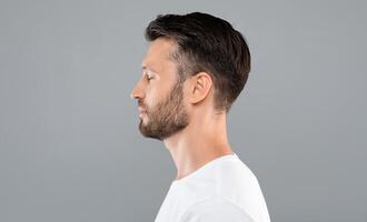 Side view of middle-aged bearded man in white t-shirt over grey studio background, copy space. Profile portrait of handsome man with closed eyes posing on gray, standing straight and looking aside photo