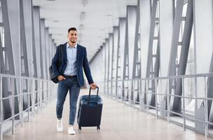 Portrait of young middle eastern man walking in airport terminal with luggage, smiling arab guy carrying suitcase while going to boarding gate, enjoying air travels and business trips, copy space photo