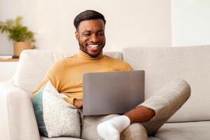 Cheerful Black Man Using Laptop Playing Computer Games Sitting On Couch At Home. Selective Focus, Copy Space photo